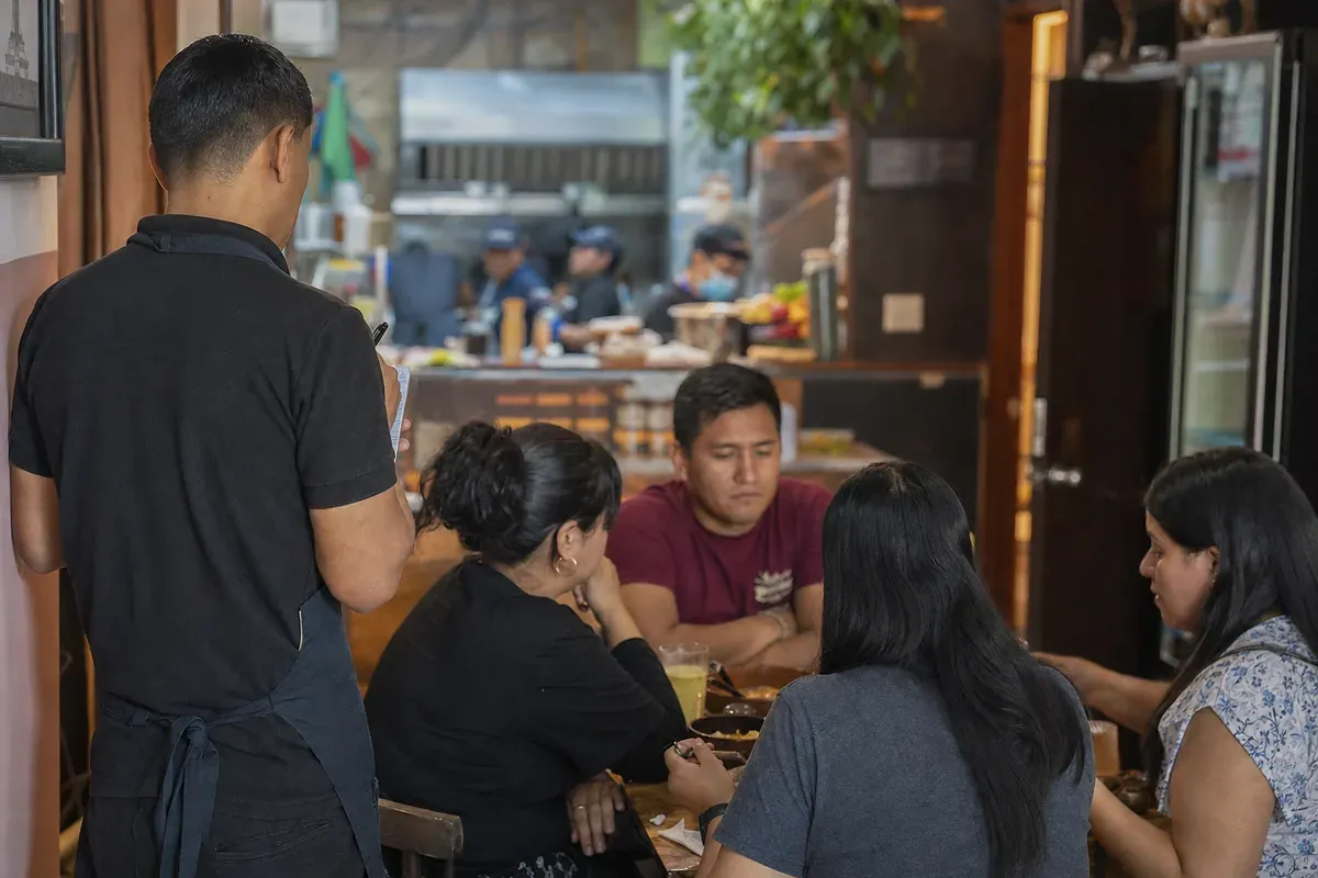 Mesero tomando el pedido en una mesa de un restaurante pequeño, con la cocina al fondo